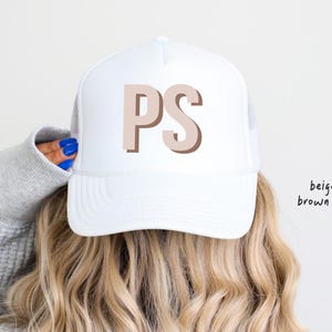 May include: A white trucker hat with the letters "PS" in beige with a brown shadow. The hat is worn by a person with long, wavy blonde hair. The text "beige top brown shadow" is in the upper right corner.