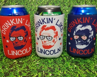 May include: Three different colored can coolers with the text "Drinkin' Like Lincoln" and a picture of Abraham Lincoln wearing sunglasses with stars on them. The can coolers are red, white, and blue.