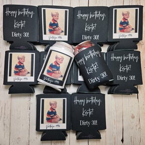 Peut inclure: Plusieurs manchons isothermes noirs pour canettes avec l'inscription blanche "Happy birthday, Kurt! Dirty 30!". Chaque manchon pr&eacute;sente une photo vintage d'un jeune gar&ccedil;on. Une canette est ins&eacute;r&eacute;e dans l'un des manchons. Photo : "Vintage 1994".