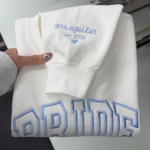 May include: A folded white sweatshirt with light blue embroidered text that reads "mrs. aguilar est. 2026" and "BRIDE". The sweatshirt is folded and held by a person. The embroidery is in a collegiate style.