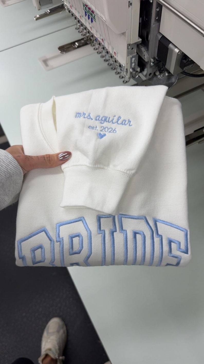 May include: A folded white sweatshirt with light blue embroidered text that reads "mrs. aguilar est. 2026" and "BRIDE". The sweatshirt is folded and held by a person. The embroidery is in a collegiate style.