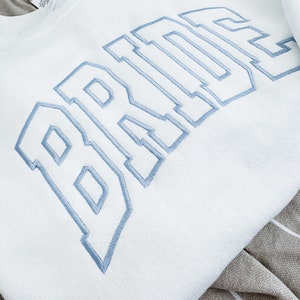 May include: White sweatshirt with the word "BRIDE" embroidered in light blue, collegiate-style lettering. The sweatshirt is folded, showing the soft texture of the fabric. The design is centered on the chest.