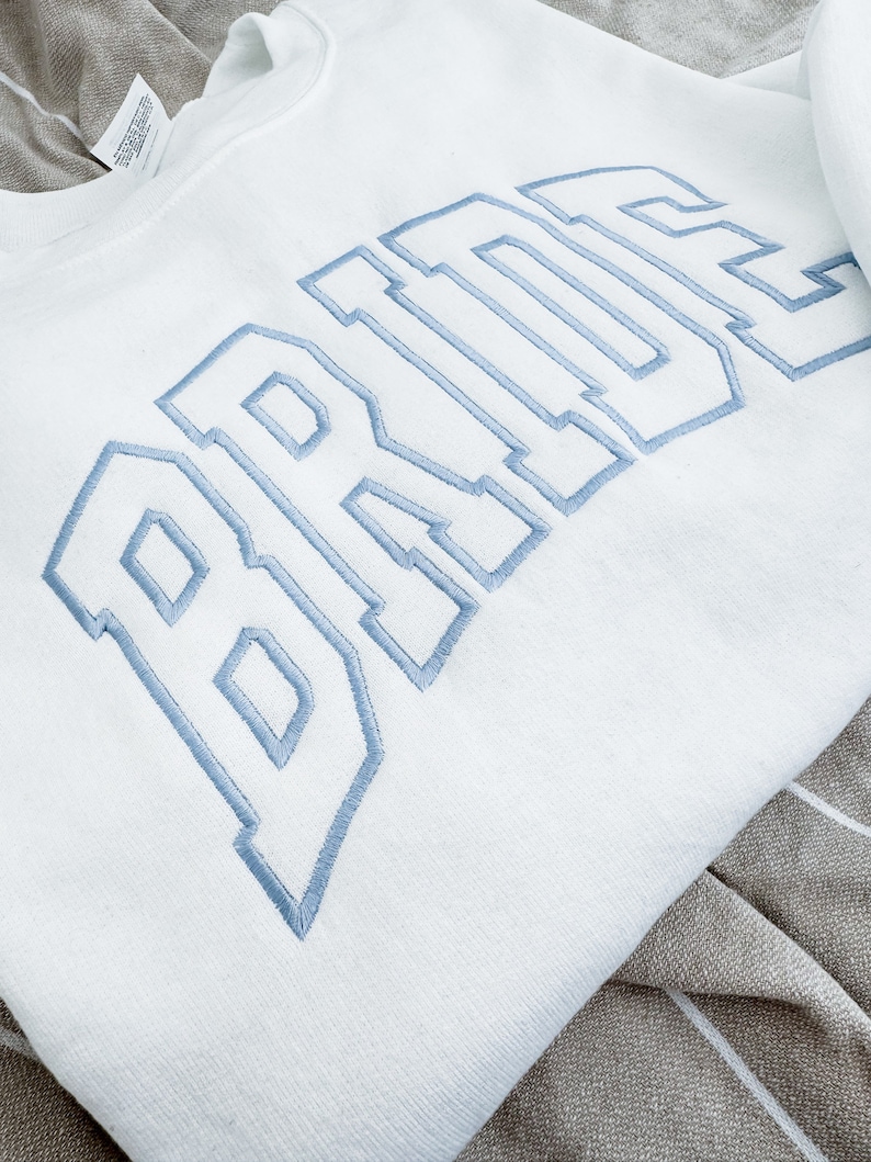 May include: White sweatshirt with the word "BRIDE" embroidered in light blue, collegiate-style lettering. The sweatshirt is folded, showing the soft texture of the fabric. The design is centered on the chest.