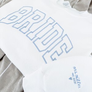May include: White sweatshirt with the word "BRIDE" embroidered in light blue, along with a matching sleeve featuring "Mrs. Miller est. 2025" in the same color. The items are displayed on a striped gray and white fabric.
