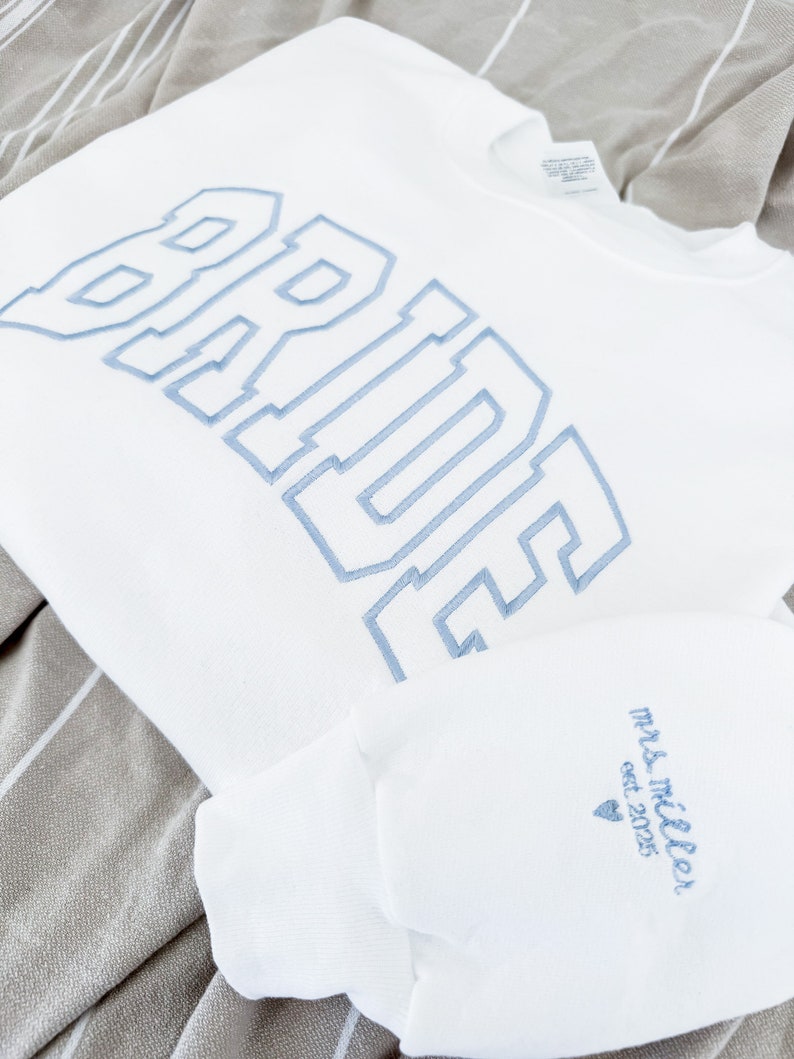 May include: White sweatshirt with the word "BRIDE" embroidered in light blue, along with a matching sleeve featuring "Mrs. Miller est. 2025" in the same color. The items are displayed on a striped gray and white fabric.