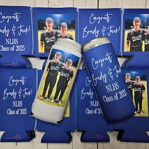 May include: Blue can coolers and koozies with the text "Congrats Brady & Jack! NLHS Class of 2025" in white script. Some coolers have a photo of two young men in baseball uniforms. The koozies are designed to keep beverages cold.