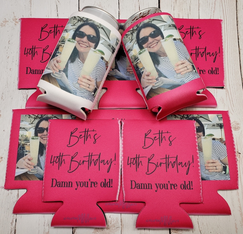 May include: Pink can coolers with the text "Beth's 40th Birthday! Damn you're old!" and a photo of a person holding a drink. The coolers are designed to keep beverages cold and are suitable for parties and celebrations.