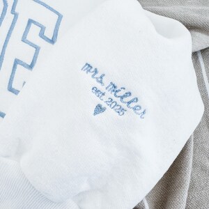 May include: White sweatshirt with light blue embroidered lettering. The sleeve features the text "mrs. miller est 2025" and a heart symbol, also in light blue. The sweatshirt is folded on a gray and white striped fabric.