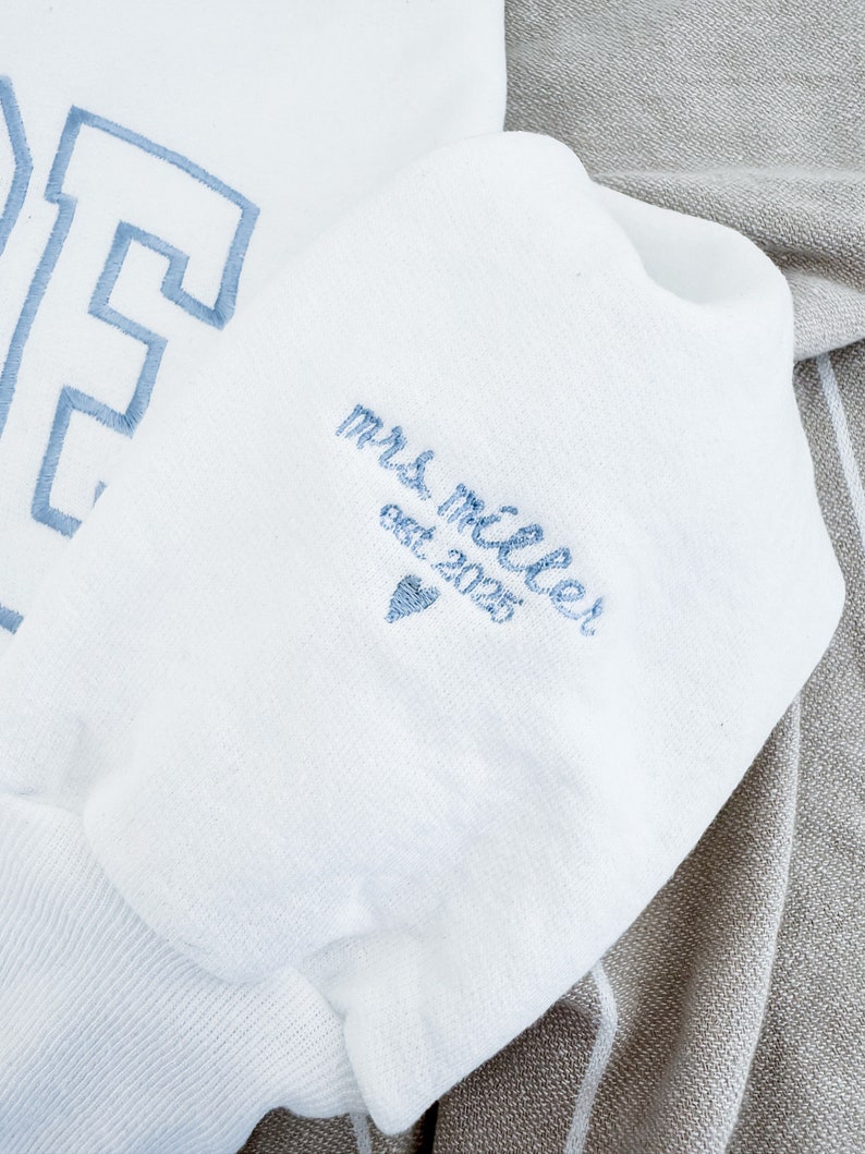 May include: White sweatshirt with light blue embroidered lettering. The sleeve features the text "mrs. miller est 2025" and a heart symbol, also in light blue. The sweatshirt is folded on a gray and white striped fabric.