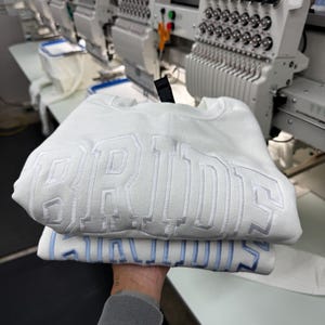May include: Two white sweatshirts with embroidered text, one stacked on top of the other. The top sweatshirt has white embroidery that reads "BRIDES". The bottom sweatshirt has light blue embroidery that reads "BRIDES". The background shows embroidery machines.