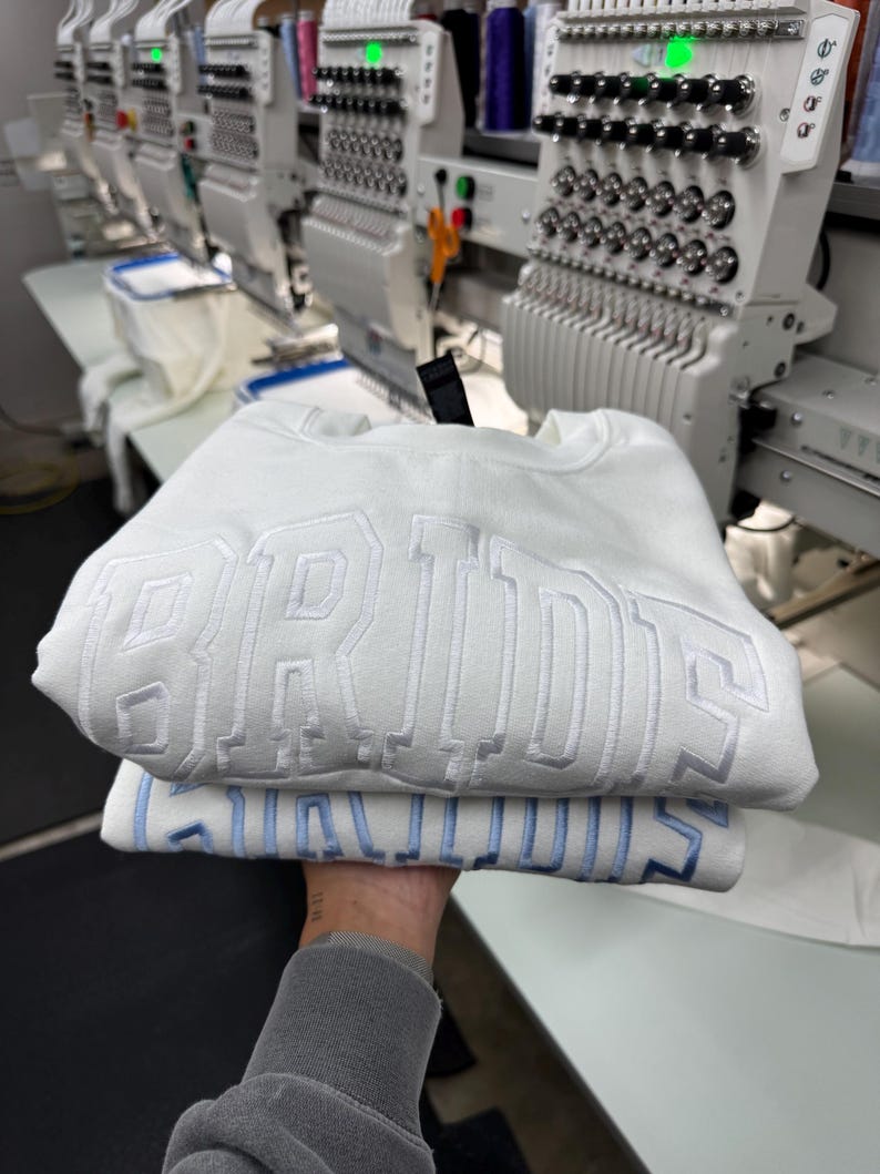 May include: Two white sweatshirts with embroidered text, one stacked on top of the other. The top sweatshirt has white embroidery that reads "BRIDES". The bottom sweatshirt has light blue embroidery that reads "BRIDES". The background shows embroidery machines.