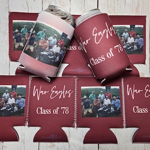 May include: Maroon can coolers with a photo of a group of people and the text "War Eagles Class of '78". One cooler is pink with a photo. The coolers are stacked on a wooden surface.