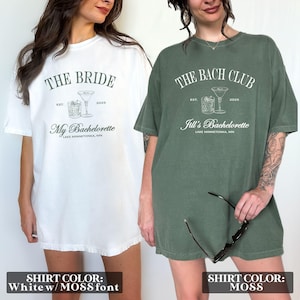 May include: Two white t-shirts with a hen party theme. The shirt on the left says "The Bride" with a cocktail glass graphic and the text "My Bachelorette" below. The shirt on the right says "The Bach Club" with a cocktail glass graphic and the text "Jill's Bachelorette" below. Both shirts have the text "EST. 2025". The shirt on the left is white with a moss green font. The shirt on the right is moss green.