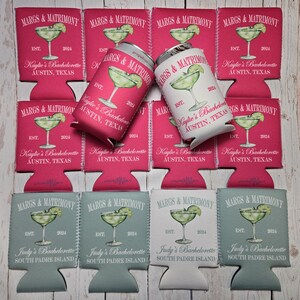 May include: A collection of drink koozies and a can, featuring a margarita design with the text "MARGS & MATRIMONY" and "Kaylie's Bachelorette Austin, Texas" or "Judy's Bachelorette South Padre Island". The koozies are pink, light blue, and white.