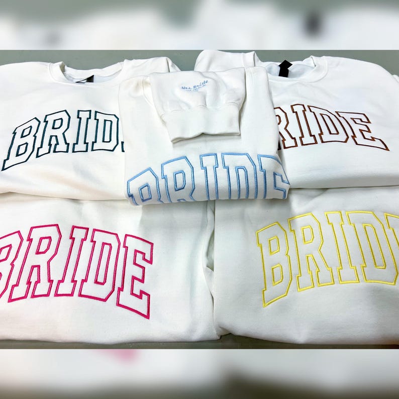 May include: White crewneck sweatshirts with the word "BRIDE" embroidered in various colors: black, blue, brown, pink, and yellow. A smaller sweatshirt with "Mrs. Bride" is also visible. The sweatshirts are likely for a bridal party.