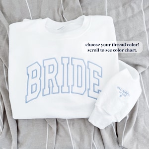 May include: White sweatshirt with the word "BRIDE" embroidered in blue on the front. The sweatshirt is folded and lying on a striped fabric. A small white tag with the text "mis nille est 2005" is attached to the sleeve.