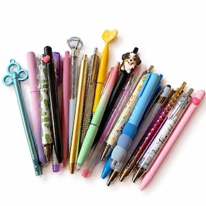 Puede incluir: Una colecci&oacute;n de bol&iacute;grafos variados con diversos dise&ntilde;os, incluyendo un bol&iacute;grafo con tope de llave azul, un tope de diamante, un tope de coraz&oacute;n amarillo, un tope de perro y varios bol&iacute;grafos con cuerpos estampados en tonos rosa, azul y oro.