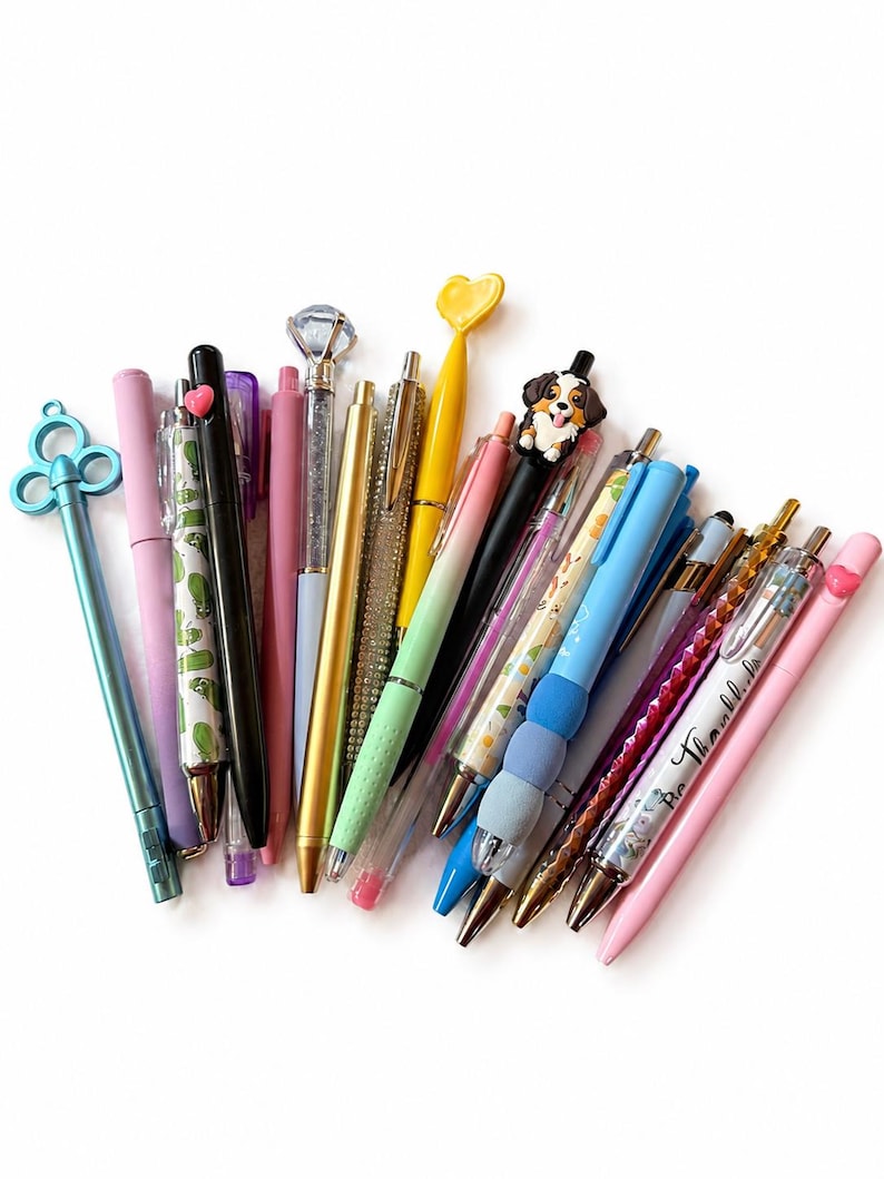 Puede incluir: Una colecci&oacute;n de bol&iacute;grafos variados con diversos dise&ntilde;os, incluyendo un bol&iacute;grafo con tope de llave azul, un tope de diamante, un tope de coraz&oacute;n amarillo, un tope de perro y varios bol&iacute;grafos con cuerpos estampados en tonos rosa, azul y oro.