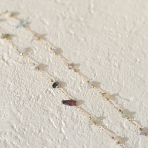 May include: Two gold chains with various colored gemstones, including blue, green, red, and black, on a white textured background.