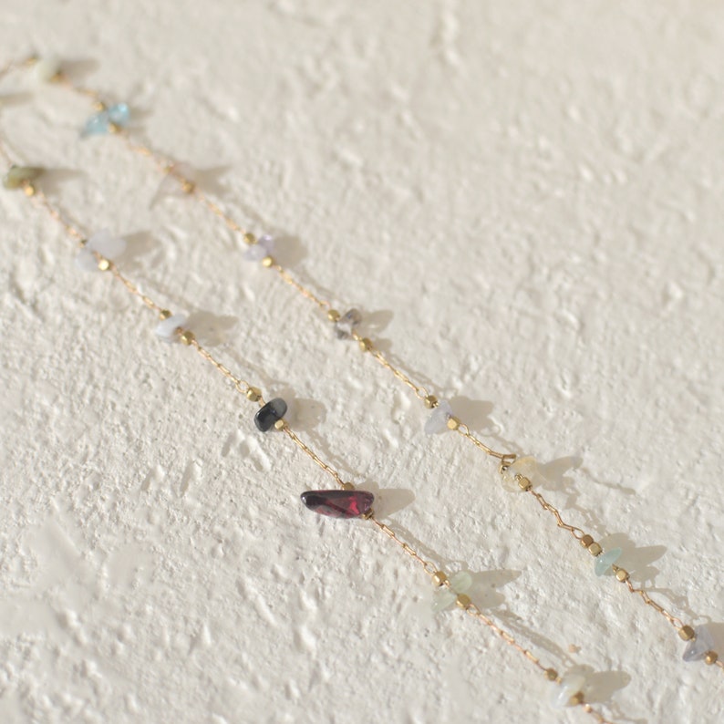 May include: Two gold chains with various colored gemstones, including blue, green, red, and black, on a white textured background.