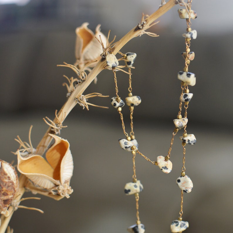 May include: A delicate gold chain necklace with black and white speckled stone beads. The necklace is hanging from a dried plant stem.