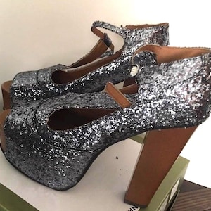 May include: A pair of silver glitter platform heels with a peep-toe design. The heels have a wooden platform and a brown ankle strap. The chunky heels are approximately 4 inches tall.