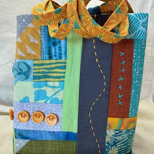 May include: A patchwork tote bag with a variety of colorful fabric panels. The bag features blue, yellow, green, and orange fabrics, with decorative stitching and buttons. The handles are a patterned orange and yellow fabric.
