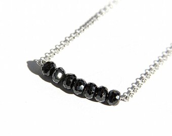 May include: A delicate necklace featuring a series of seven faceted black beads strung on a fine silver chain. The beads are evenly spaced, creating a linear design. The necklace is displayed against a bright white background.