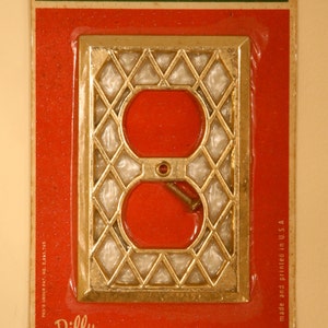Puede incluir: Una tapa de toma de corriente de color dorado con un patrón de diamante. La tapa está empaquetada en una caja roja y verde con el texto "decro plate elegance in your home" y "Dilly MFG. CO., INC., S. PLAINFIELD, N. J. 07080 made and printed in U.S.A."