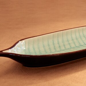 May include: A long, narrow ceramic serving dish shaped like a leaf. The dish is glazed in a light green color with a brown rim.