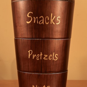 May include: A brown wooden tiered container with a lid. The container has three sections labeled "Snacks", "Pretzels", and "Nuts".