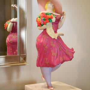 May include: A decorative figurine of a woman in a pink dress and large hat, holding a bouquet of flowers. The dress has a gold floral pattern. The figurine stands on a white marble base. A mirror reflects the figurine.