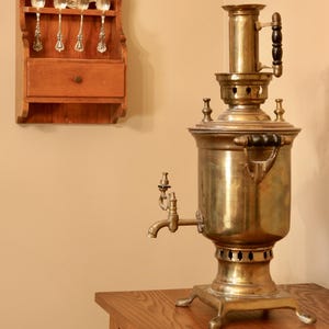 May include: A vintage brass samovar with a spigot and black handles sits on a wooden table. A wooden spoon rack with ornate silver spoons hangs on the wall. The scene evokes a sense of traditional tea service.