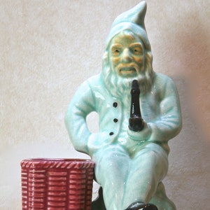 Puede incluir: Una figurita de cerámica de un gnomo sentado en una pequeña maceta de mimbre roja. El gnomo lleva un atuendo azul claro y tiene una larga barba blanca. Sostiene una pipa negra en su mano derecha.