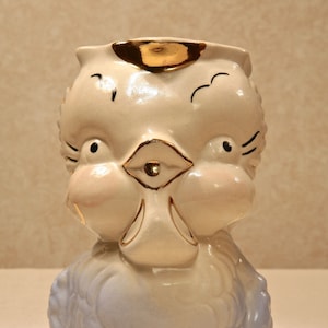 Puede incluir: Un pichel de cerámica vintage con forma de pájaro con un esmalte azul y blanco. El pájaro tiene un borde dorado y detalles pintados en su rostro.