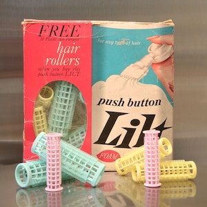 May include: A box of hair rollers with the text 'FREE 36 Plastic All Purpose hair rollers when you buy this push button LILT' and 'push button Lil FOAM' on it. The box is pink and blue with a picture of a woman using a hair product. There are several hair rollers in front of the box, some are pink, some are yellow, and some are green.