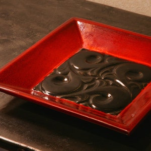Puede incluir: Un plato cuadrado de cerámica roja con un diseño negro, en relieve y complejo en el centro.