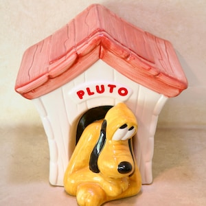 May include: A ceramic cookie jar shaped like a doghouse with a red roof and white walls. The word PLUTO is printed in red letters above the entrance. A yellow dog with black ears and a black nose is positioned in the front of the doghouse entrance.