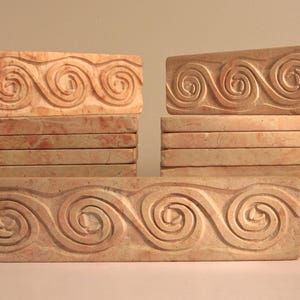 May include: A set of six rectangular tiles with a carved design of swirling waves. The tiles are made of a light brown stone with a reddish hue.