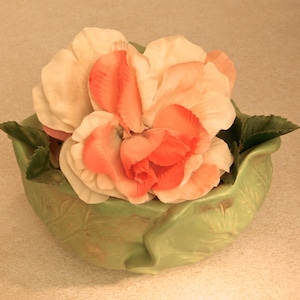 Può includere: Composizione decorativa con grandi fiori a strati nei toni del pesca e del corallo, adagiati in una ciotola di ceramica verde chiaro a forma di foglia. La ciotola ha una superficie strutturata ed è riempita di fiori artificiali.