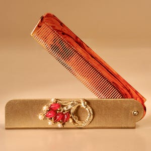 May include: A vintage folding comb with a tortoiseshell pattern comb and a gold-toned case. The case is decorated with red jewels and small white pearls. The comb is partially open, revealing the fine teeth.