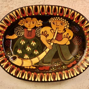 May include: Oval ceramic platter with a black background and a decorative border. The platter features a folk art design of two figures, one playing a horn. The figures are in a traditional style with bright colors.