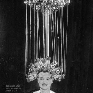 May include: A woman with her hair in curlers under a metal hair dryer with multiple bulbs. The hair dryer is suspended from the ceiling. The woman is wearing a white lab coat with a black badge that says 'ICAL'.
