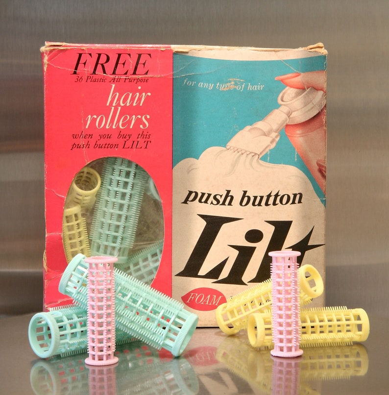 May include: A box of hair rollers with the text 'FREE 36 Plastic All Purpose hair rollers when you buy this push button LILT' and 'push button Lil FOAM' on it. The box is pink and blue with a picture of a woman using a hair product. There are several hair rollers in front of the box, some are pink, some are yellow, and some are green.