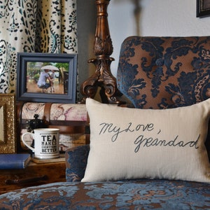 Peut inclure: Un coussin blanc avec le texte "My love, Granddad" brodé en écriture cursive noire. Le coussin est posé sur un fauteuil à motifs marrons et bleus.