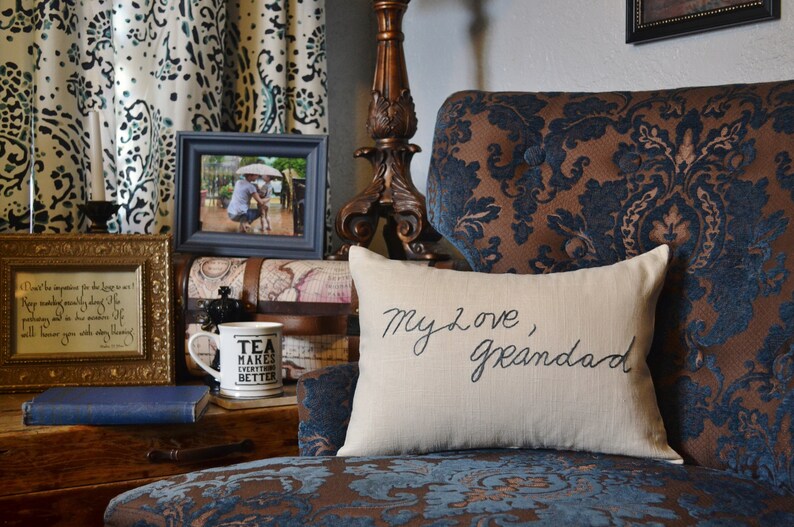 Puede incluir: Una almohada blanca con el texto "My love, Granddad" bordado en letra cursiva negra. La almohada est&aacute; sentada en un sill&oacute;n de estampado marr&oacute;n y azul.