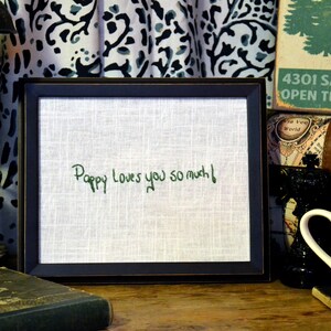 May include: A framed piece of fabric with the words "Pappy Loves you so much!" embroidered in green thread. The frame is black and has a white mat.