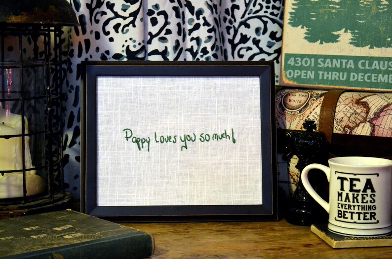 May include: A framed piece of fabric with the words "Pappy Loves you so much!" embroidered in green thread. The frame is black and has a white mat.