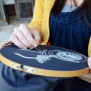 May include: A person is embroidering a white bicycle design on a black fabric stretched in a wooden embroidery hoop. The hoop is held in the person's hands. The person is wearing a yellow cardigan.