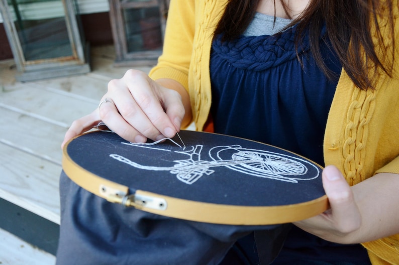 May include: A person is embroidering a white bicycle design on a black fabric stretched in a wooden embroidery hoop. The hoop is held in the person's hands. The person is wearing a yellow cardigan.
